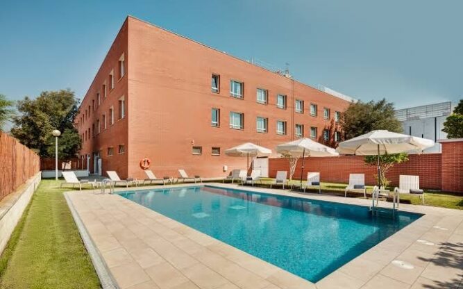 Exterior of Sercotel, with a large pool in front of the building and blue skies above.