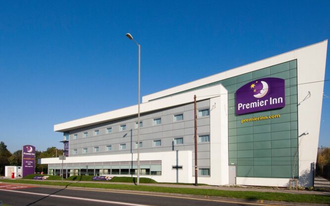 The front view of Premier Inn Liverpool Airport Hotel, a white building with lots of windows and a purple hotel sign.