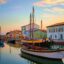 A traditional Italian boat sat on the canal in Cesenatico with houses along the shore and the sky painted in the pastel colours of dusk.