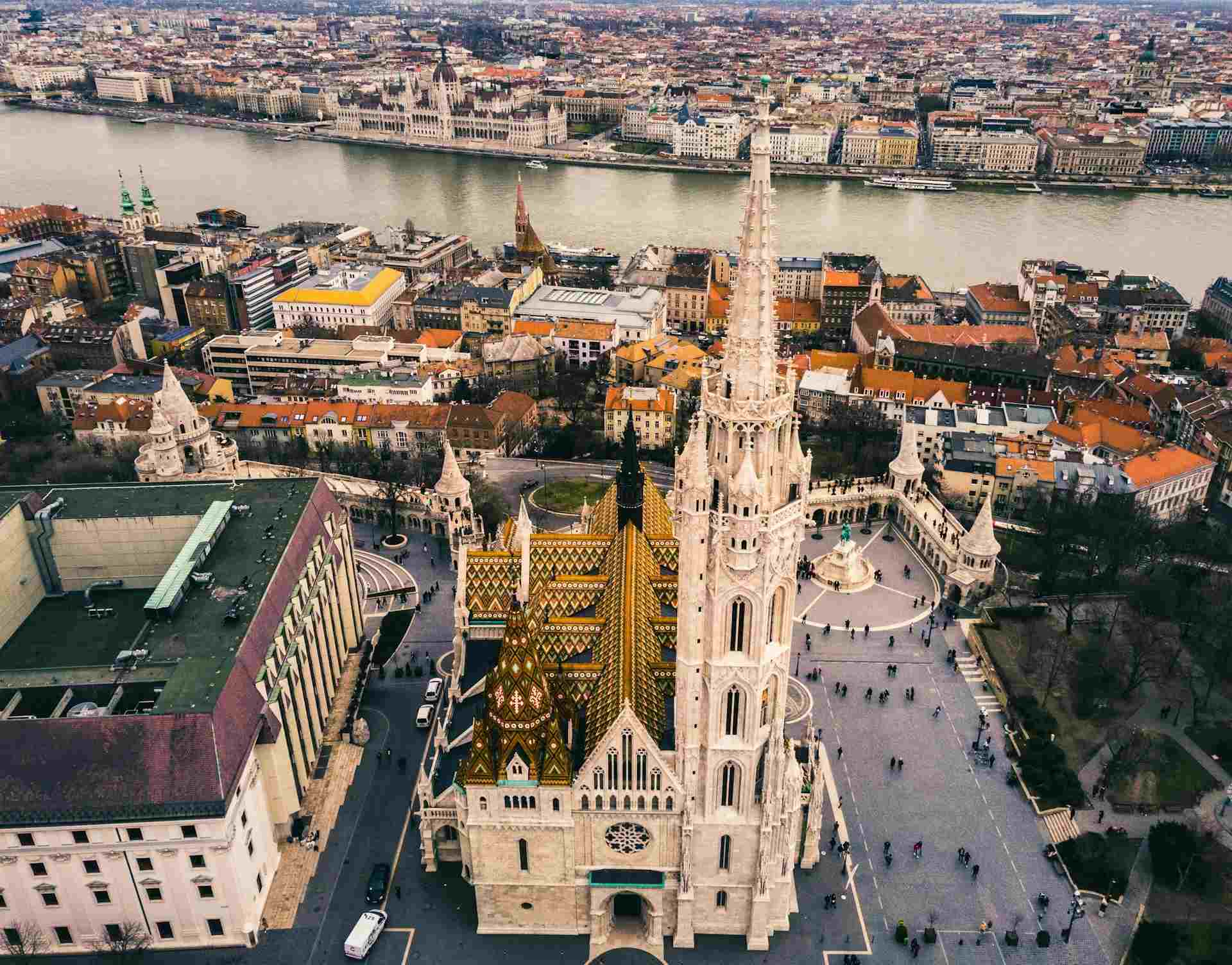 A birds eye view of central Budapest, with Matthias Church in the center and traditional buildings surrounding it., as well as a river running running through the center.
