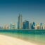 View of Abu Dhabi’s modern skyline with glass skyscrapers and clear blue waters seen from a sandy beach on a bright day, representing the final destination for travelers journeying from Dubai International Airport to Abu Dhabi.