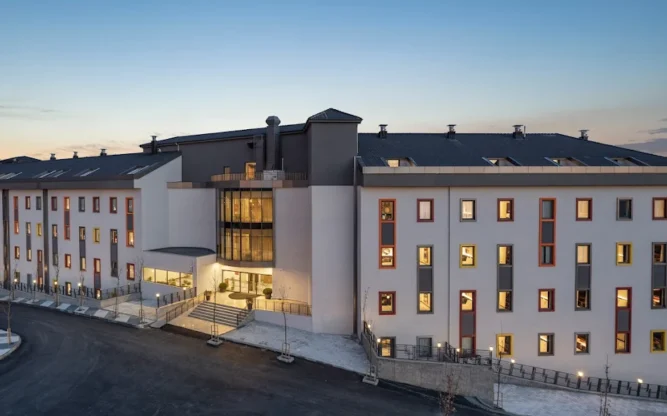 Exterior view of Hampton by Hilton near Istanbul Airport at dusk, featuring a modern multi-story building with illuminated windows, landscaped pathways, and a quiet, welcoming atmosphere against a soft evening sky.