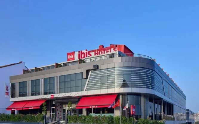 Modern exterior of the Ibis Istanbul Airport hotel during daytime, featuring large glass windows, red awnings, and a sleek curved facade, conveniently located near Istanbul Airport.