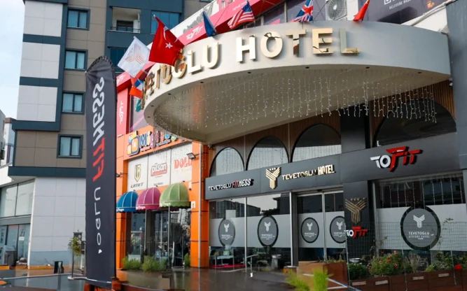 Exterior view of Tevetoglu Hotel during daytime, featuring a curved entrance canopy, multiple flags, and gym signage—showcasing the modern facade of this popular stay near Istanbul Airport.