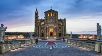 Evening view of the Ta’ Pinu Basilica in Gozo, illuminated against a cloudy sky, with statues lining the entrance path — a popular landmark often visited by travelers going to Gozo from Malta Airport.