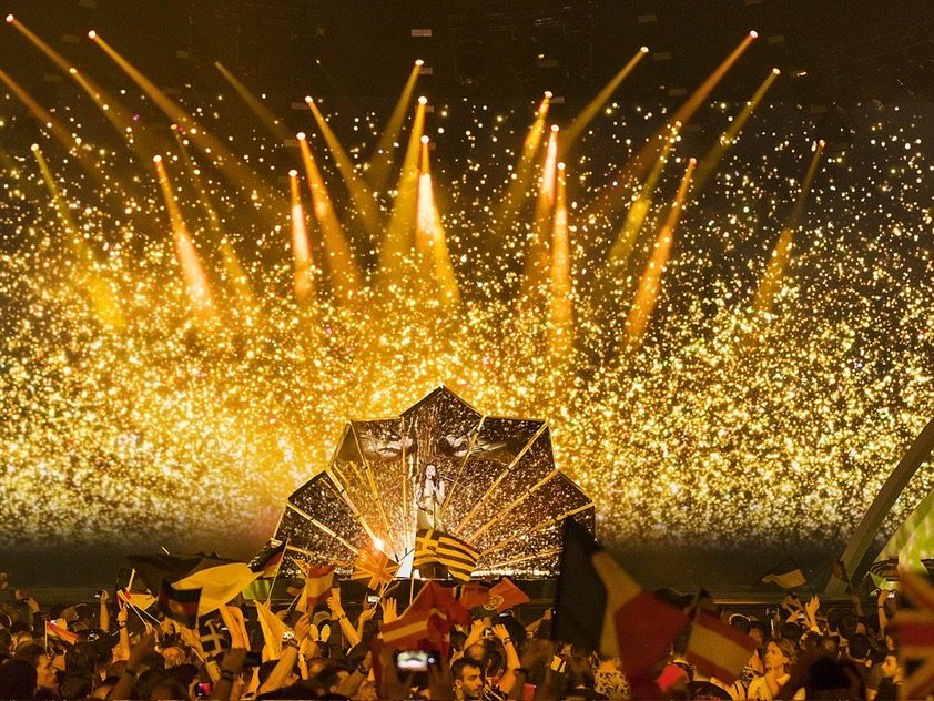 Vibrant Eurovision 2026 Grand Final stage with a single performer on a mirrored platform, dramatic gold lighting, and the audience waving national flags.