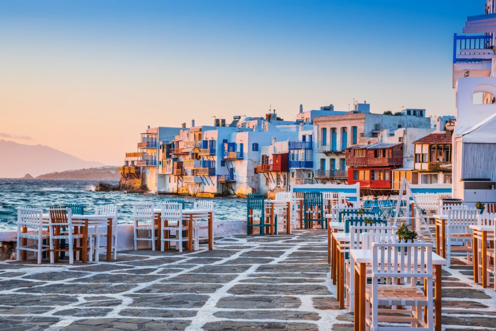 
"Little Venice in Mykonos at sunset, whitewashed and colorful neoclassical houses built directly on the rocks above the turquoise Aegean Sea, with a stone-paved waterfront promenade lined with white and blue outdoor dining tables and chairs, beneath a warm sky glowing in shades of orange and blue."