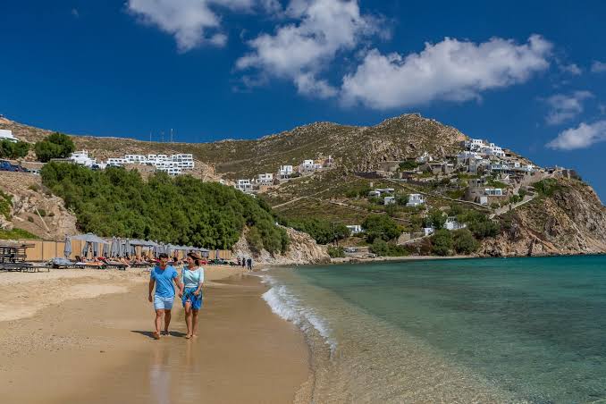 Scenic view of a Mykonos beach with turquoise Aegean waters, golden sand, and white buildings built into the rocky hills under a bright blue sky.