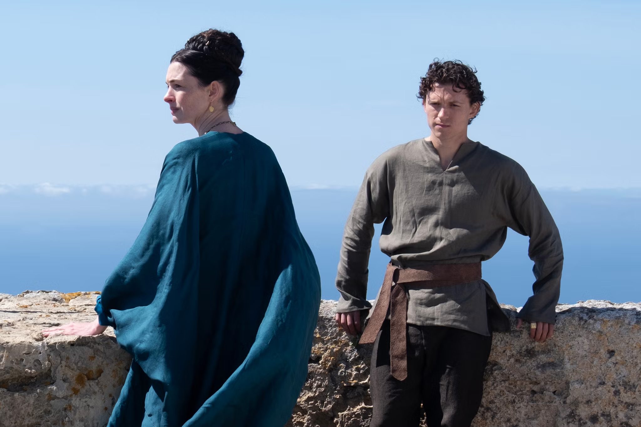 Anne Hathaway as Penelope and Tom Holland as Telemachus standing by a stone wall overlooking the ocean in Christopher Nolan's 2026 film, The Odyssey.