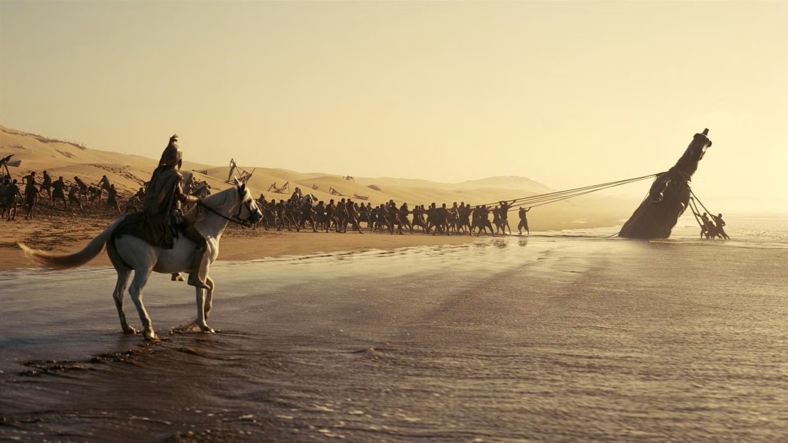 A Greek warrior on a white horse watching an army pull a massive wooden ship onto a sandy beach in a cinematic scene from Christopher Nolan's 2026 film, The Odyssey.