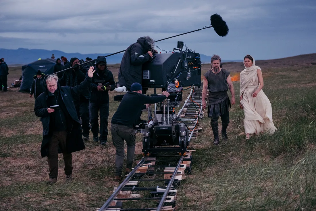 Director Christopher Nolan on the outdoor set of The Odyssey (2026) with an IMAX camera crew, as stars Matt Damon and Zendaya walk in character behind them.