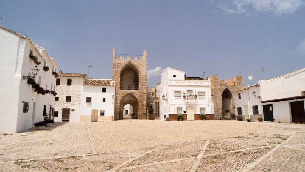 Wide view of a paved cobblestone plaza flanked by white Mediterranean buildings and historic stone fortified towers with arched gateways under a clear sky.